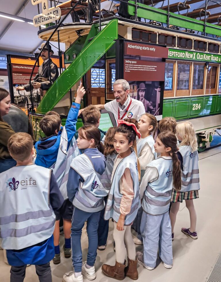 EIFA A group of children in reflective vests gather around an older man in front of a vintage green tram inside a museum, listening attentively as one child raises their hand.