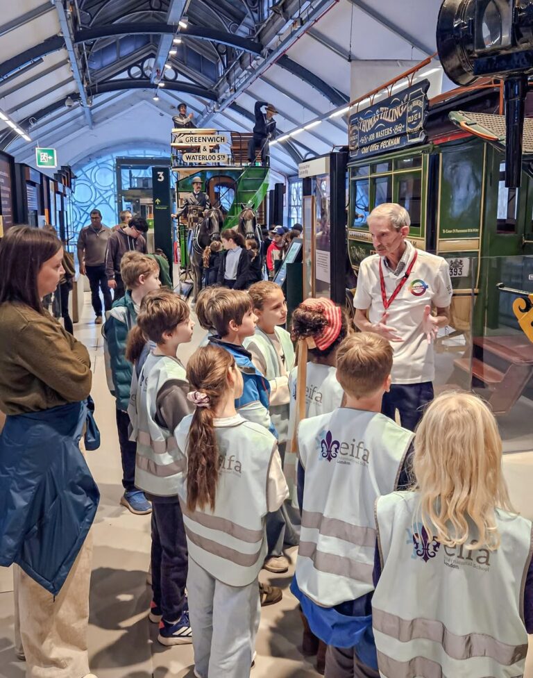 EIFA A group of children in matching vests listens to a museum guide explaining an exhibit in a transport museum, with vintage vehicles and signs visible in the background. An adult stands nearby, observing the group.