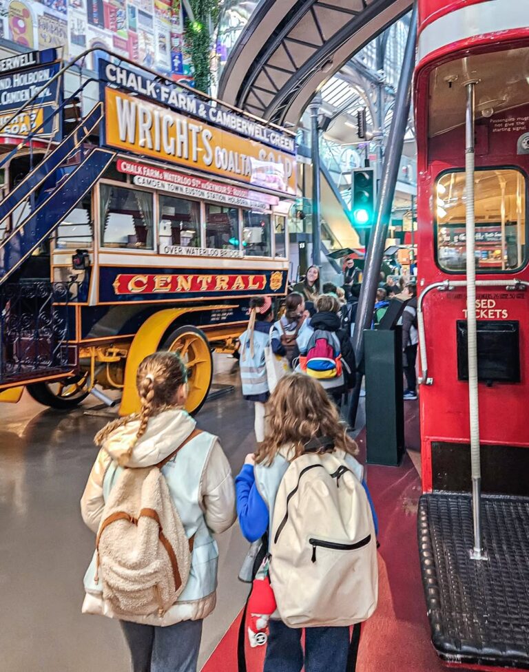 EIFA Children with backpacks walk inside a museum, passing by a vintage double-decker bus and a historic horse-drawn carriage with colorful signage, surrounded by displays and bright lights.