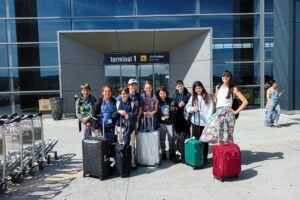 EIFA A group of eight children and one adult stand outside an airport terminal with their suitcases, smiling at the camera during International Open Day. The entrance behind them reads terminal 1 Arrivées / Arrivals. EIFA A group of eight children and one adult stand outside an airport terminal with their suitcases, smiling at the camera during International Open Day. The entrance behind them reads terminal 1 Arrivées / Arrivals.