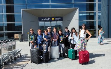 EIFA A group of eight children and one adult stand outside an airport terminal with their suitcases, smiling at the camera during International Open Day. The entrance behind them reads terminal 1 Arrivées / Arrivals.