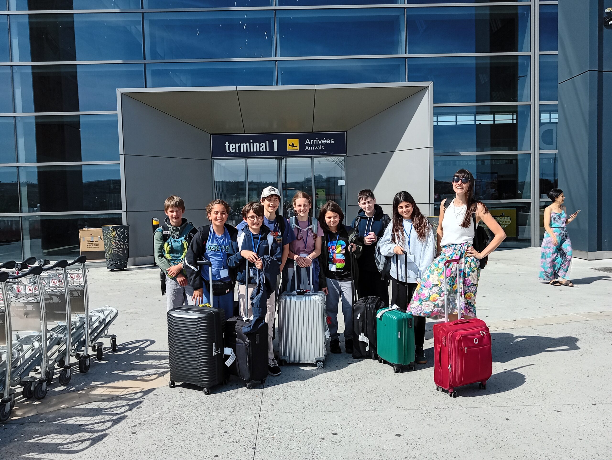 EIFA A group of eight children and one adult stand outside an airport terminal with their suitcases, smiling at the camera during International Open Day. The entrance behind them reads terminal 1 Arrivées / Arrivals.