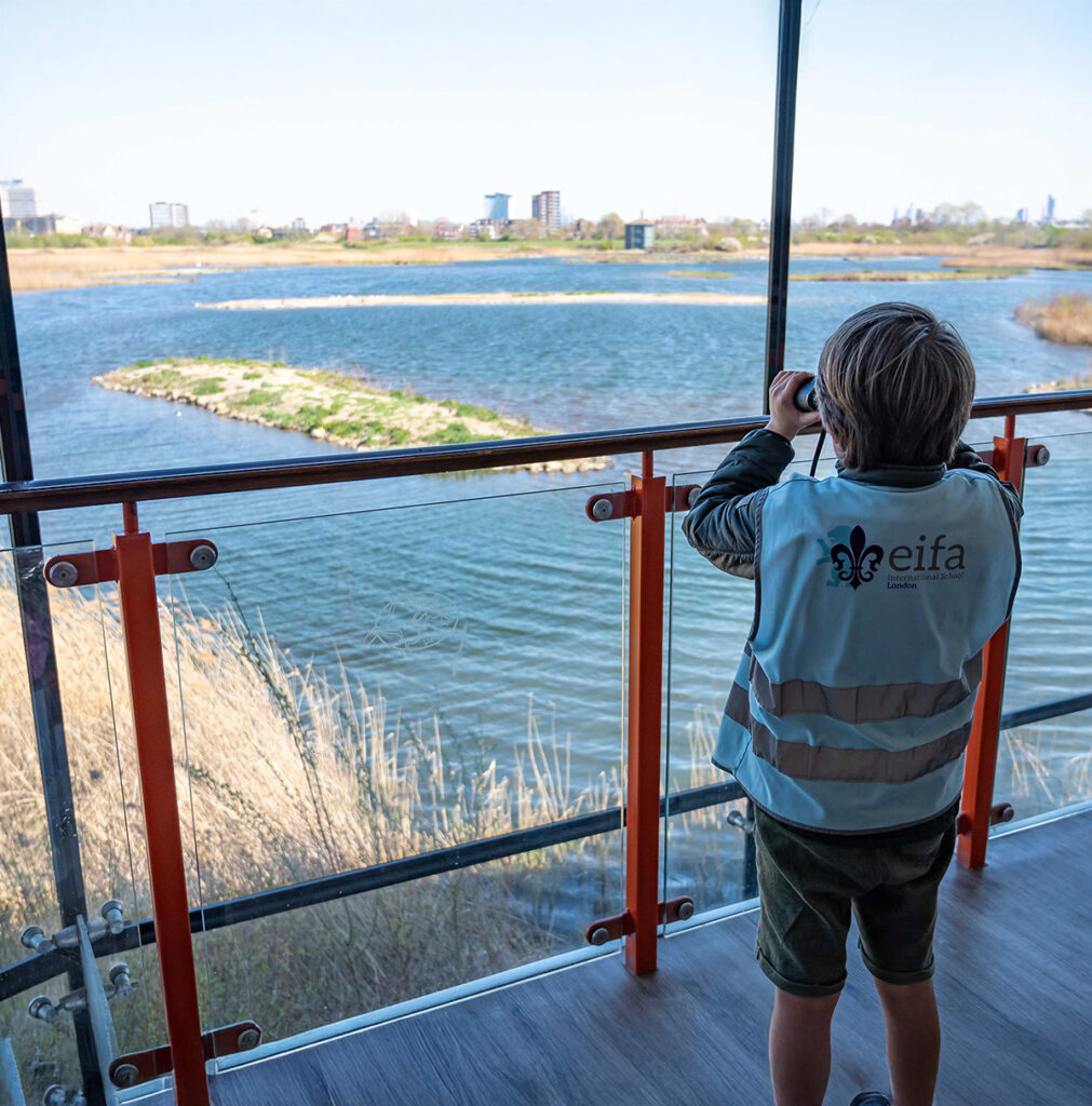 EIFA A child wearing a light blue vest is looking through binoculars at a large lake from a glass balcony. The scene outside includes grassy land and a few tall buildings in the distance under a clear blue sky.