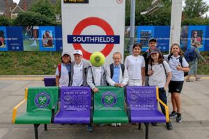 EIFA Un groupe d'enfants souriants, vêtus de tenues de sport, posent à la gare de Southfields. Des banderoles aux couleurs de Wimbledon et une balle de tennis sont disposées sur les bancs, créant une ambiance festive à l'occasion de la Journée mondiale de l'alimentation. Des pancartes et de la verdure en arrière-plan viennent rehausser cette scène pleine de vie.