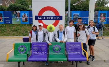 EIFA A group of kids in sportswear pose at Southfields Underground station with tennis props and Wimbledon banners on benches behind them, celebrating International Open Day.