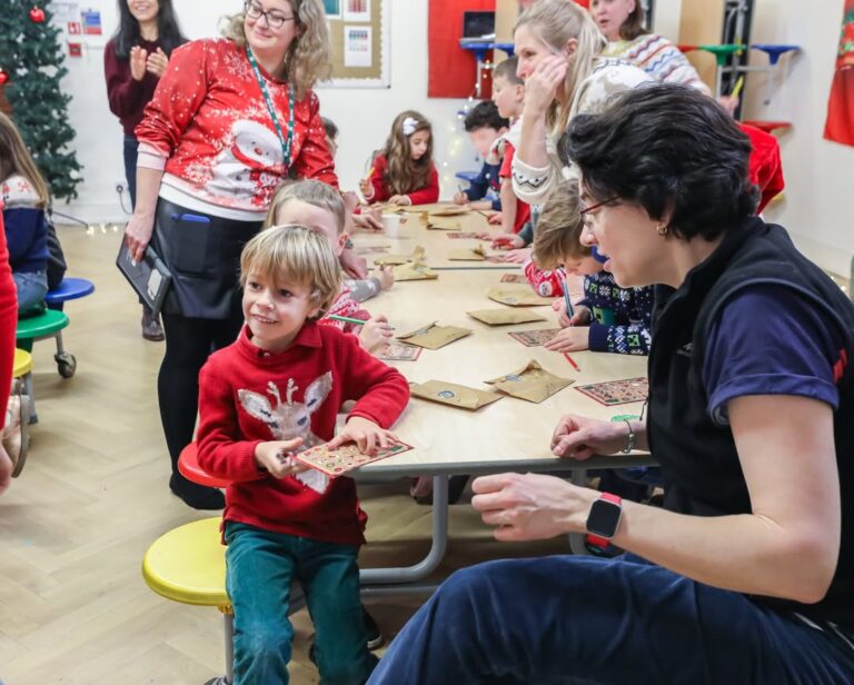 EIFA Children and adults sit around a table in a festive classroom, making crafts for International Open Day. A smiling boy in a red reindeer sweater holds a decorated item while talking to an adult. Holiday decorations brighten the background.