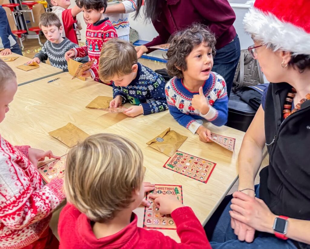 EIFA Des enfants vêtus de pulls festifs jouent au bingo sur le thème de Noël autour d'une table, sous la surveillance d'une femme coiffée d'un bonnet de Père Noël. Des sacs en papier marron marquent l'occasion, ajoutant à l'atmosphère animée inspirée par la Journée mondiale de l'alimentation.