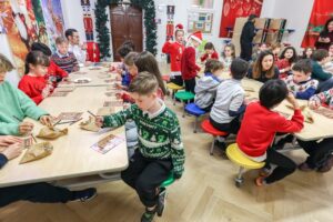 EIFA Des enfants sont assis autour de tables dans une salle de classe festive, coloriant sur des sacs en papier dans le cadre d’une activité de la Journée mondiale de l’alimentation. Des décorations de vacances, notamment des couronnes et des affiches sur le thème de Noël, ornent la salle tandis que des adultes supervisent et assistent les enfants. EIFA Des enfants sont assis autour de tables dans une salle de classe festive, coloriant sur des sacs en papier dans le cadre d'une activité de la Journée mondiale de l'alimentation. Des décorations de vacances, notamment des couronnes et des affiches sur le thème de Noël, ornent la salle tandis que des adultes supervisent et assistent les enfants.