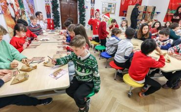 EIFA A group of children sit around tables in a festive classroom during International Open Day, wearing colorful sweaters. They draw or write on paper under adult supervision, surrounded by Christmas decorations and artwork on the walls.