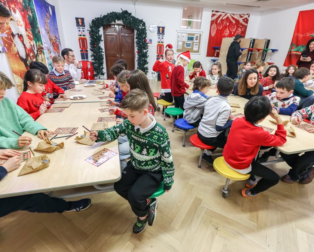 EIFA Des enfants sont assis autour de tables dans une salle de classe festive, coloriant sur des sacs en papier dans le cadre d'une activité de la Journée mondiale de l'alimentation. Des décorations de vacances, notamment des couronnes et des affiches sur le thème de Noël, ornent la salle tandis que des adultes supervisent et assistent les enfants.