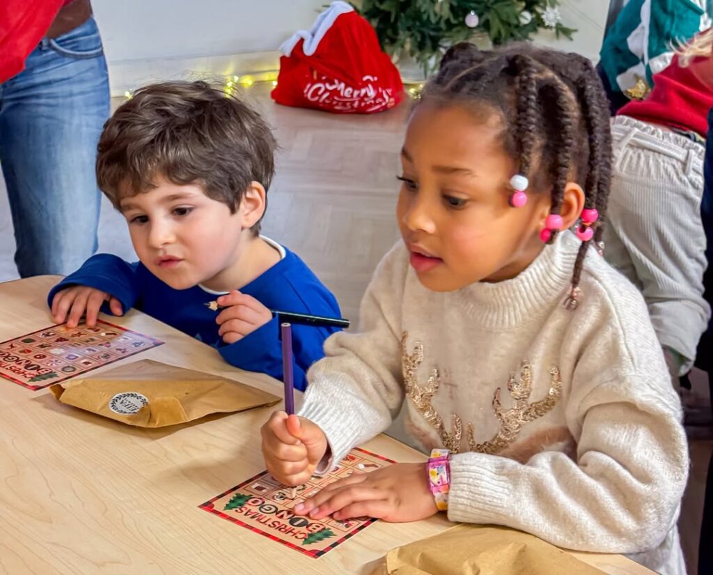 EIFA Deux jeunes enfants sont assis à une table, coloriant des feuilles d'activités sur le thème des fêtes, à l'occasion de la Journée mondiale de l'alimentation. La fille en pull blanc tient un stylo tandis que le garçon en bleu regarde, avec des décorations de vacances et un arbre de Noël derrière eux.