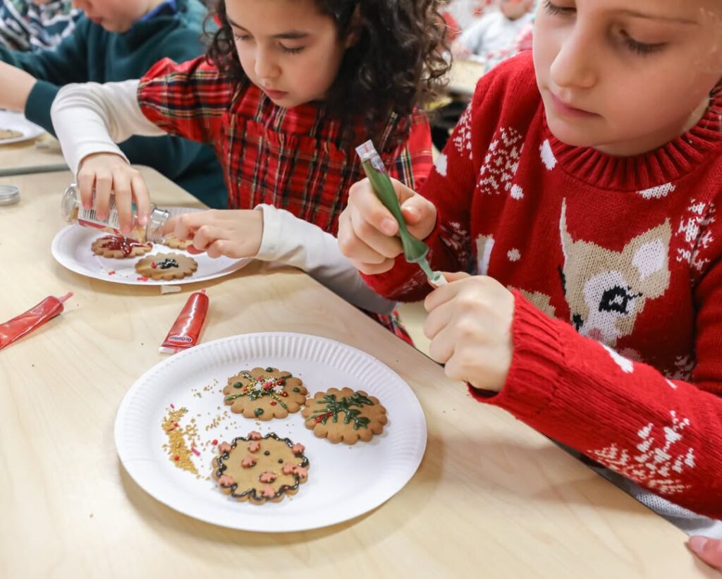 EIFA Deux enfants décorent des biscuits de Noël avec du glaçage et des paillettes à une table, célébrant ainsi la créativité et la convivialité - parfait pour la Journée mondiale de l'alimentation. Les biscuits décorés sont posés sur des assiettes en papier blanc devant eux.