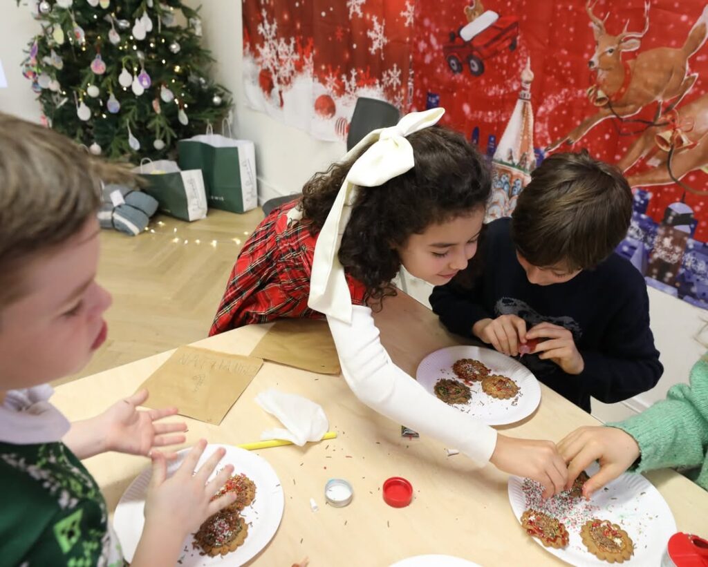 EIFA Trois enfants décorent des biscuits avec des paillettes sur une table. Un arbre de Noël et des décorations festives créent une ambiance de vacances, tandis que les enfants s'adonnent à leur activité - parfaite pour célébrer la convivialité lors de la Journée mondiale de l'alimentation.