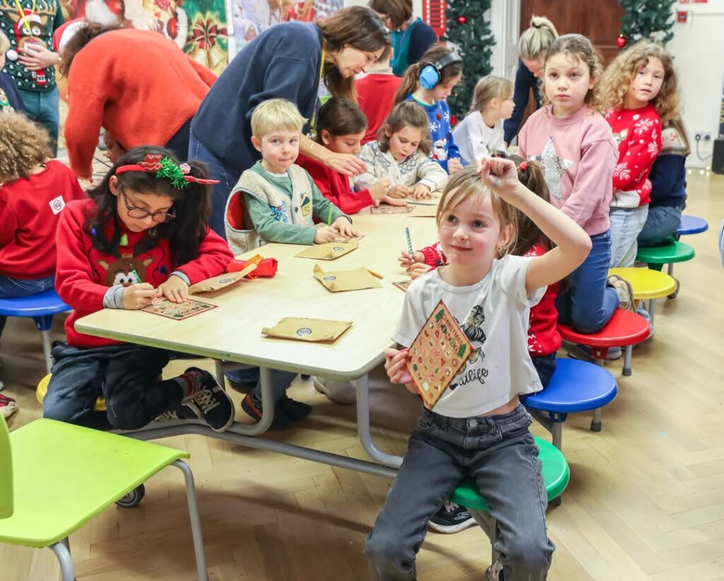 EIFA Un groupe de jeunes enfants est assis à des tables colorées et participe à une activité artisanale festive dans le cadre de la Journée mondiale de l'alimentation. L'un d'entre eux brandit sa carte décorée et sourit, tandis que des adultes l'assistent dans la salle lumineuse décorée pour les fêtes.