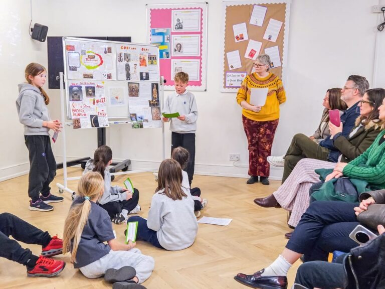 EIFA Two children stand by a display board presenting to a seated audience of adults and children. An adult woman stands nearby, observing and smiling. Classroom setting with colorful posters on the wall.