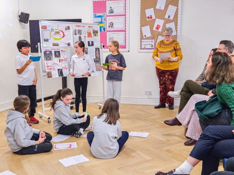 EIFA A group of children present and read from papers in a classroom; some stand near a whiteboard covered in images and notes, while others sit on the floor. Adults sit watching; a woman stands, smiling in the background.