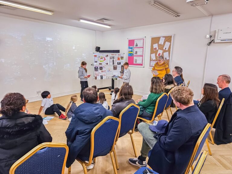 EIFA Children present a project in front of a display board to an audience of adults and kids seated on blue chairs in a classroom with bright lighting and posters on the walls.