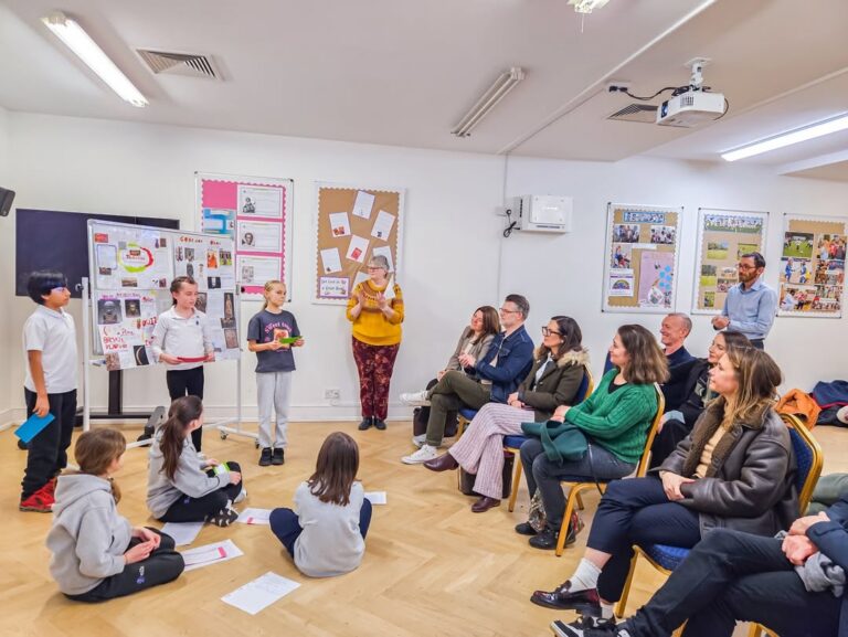 EIFA Children give a presentation to seated adults in a classroom. Some kids stand near a display board, while others sit on the floor. The adults watch attentively. Educational posters and papers decorate the walls.
