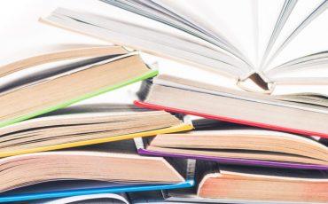 EIFA A vibrant stack of open books, reminiscent of a cozy Soho nook, is set in two rows with their pages artfully fanned out. The colorful covers, in shades like blue and green, invite readers to discover new stories against a plain white backdrop.