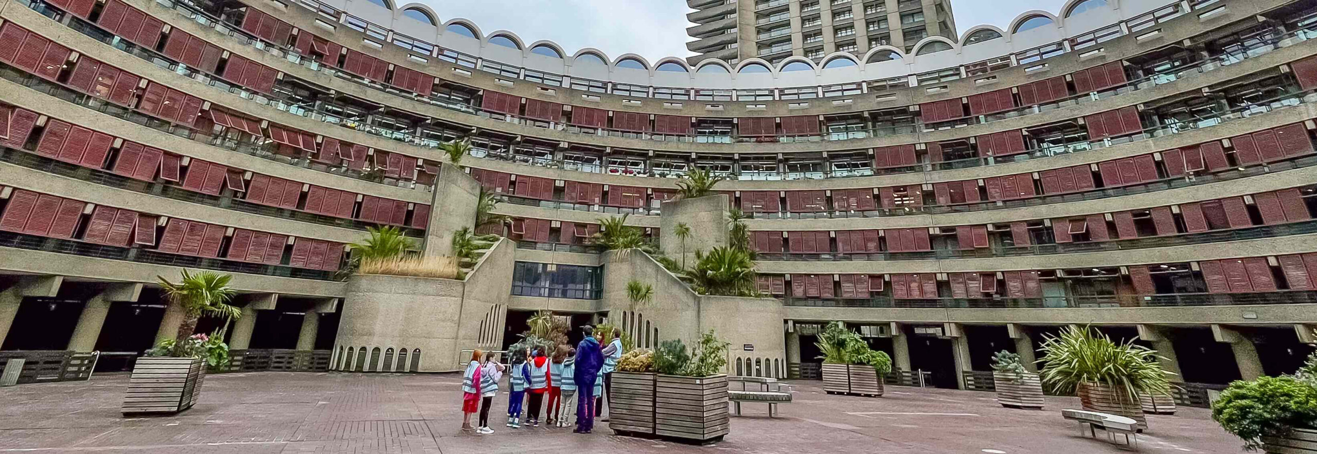 EIFA A group of people stands in the large, circular courtyard of a modern bilingual school in London, featuring multiple floors and red window shutters. Potted plants are scattered around the paved open area, and a tall tower looms in the background.