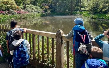 EIFA Un groupe d'enfants avec des sacs à dos et deux adultes se tiennent près d'une clôture en bois au London Wetland Centre, admirant un étang entouré de verdure par une journée ensoleillée. Un adulte pointe du doigt quelque chose au loin pendant leur visite au London Wetland Centre.