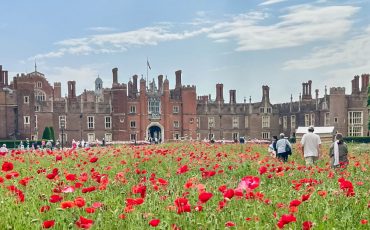 EIFA Year 3 and Year 4 visited the Isle of Wight, where people walk through a field of red poppies in front of a large historic brick building with many chimneys and turrets under a partly cloudy sky, surrounded by trees and groups of tourists.