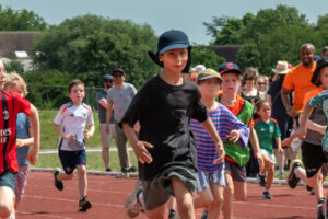 EIFA Reception pupils at Hampton Court Palace race on a running track during a sports event, with spectators and adults cheering enthusiastically in the background on a sunny day.