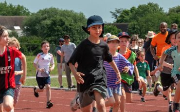 EIFA Reception pupils at Hampton Court Palace race on a running track during a sports event, with spectators and adults cheering enthusiastically in the background on a sunny day.