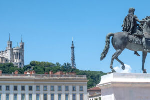 EIFA La statue d'un homme à cheval se dresse sur une place de la ville entourée de bâtiments classiques ; à l'arrière-plan, la colline abrite la basilique Notre-Dame de Fourvière et une haute tour métallique, plantant le décor d'une Journée des sports animée sous un ciel bleu limpide.