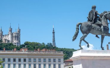 EIFA La statue d'un homme à cheval se dresse sur une place de la ville entourée de bâtiments classiques ; à l'arrière-plan, la colline abrite la basilique Notre-Dame de Fourvière et une haute tour métallique, plantant le décor d'une Journée des sports animée sous un ciel bleu limpide.