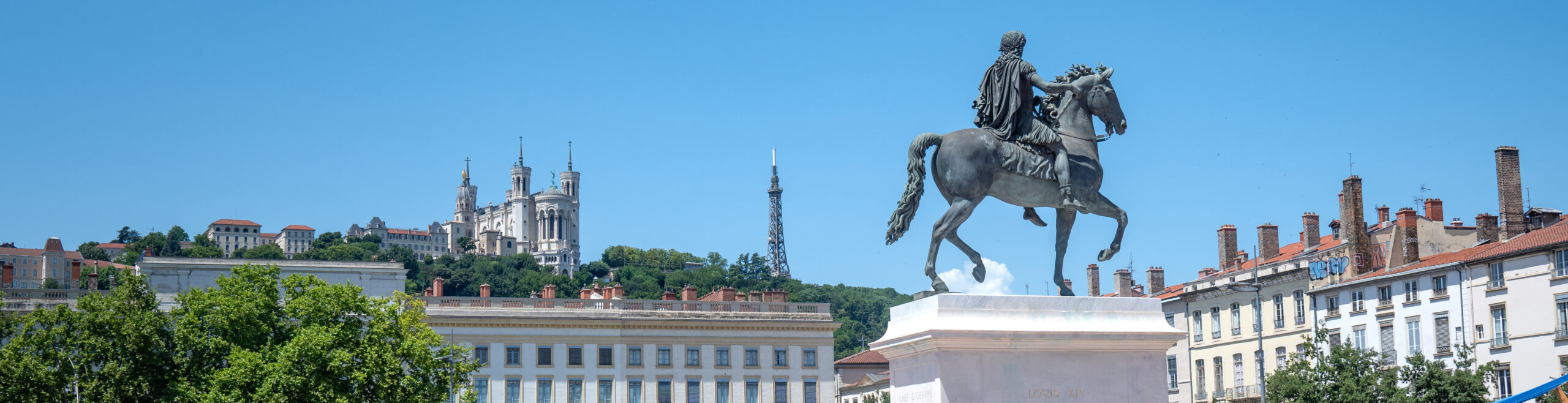 EIFA La statue d'un homme à cheval se dresse sur une place de la ville entourée de bâtiments classiques ; à l'arrière-plan, la colline abrite la basilique Notre-Dame de Fourvière et une haute tour métallique, plantant le décor d'une Journée des sports animée sous un ciel bleu limpide.
