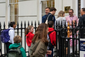 EIFA A group of parents and children with backpacks, including Year 5 and 6 students heading to Lyon, walk toward the entrance of a city building, where several adults are cheerfully greeting them—capturing the bustle of a lively school drop-off.