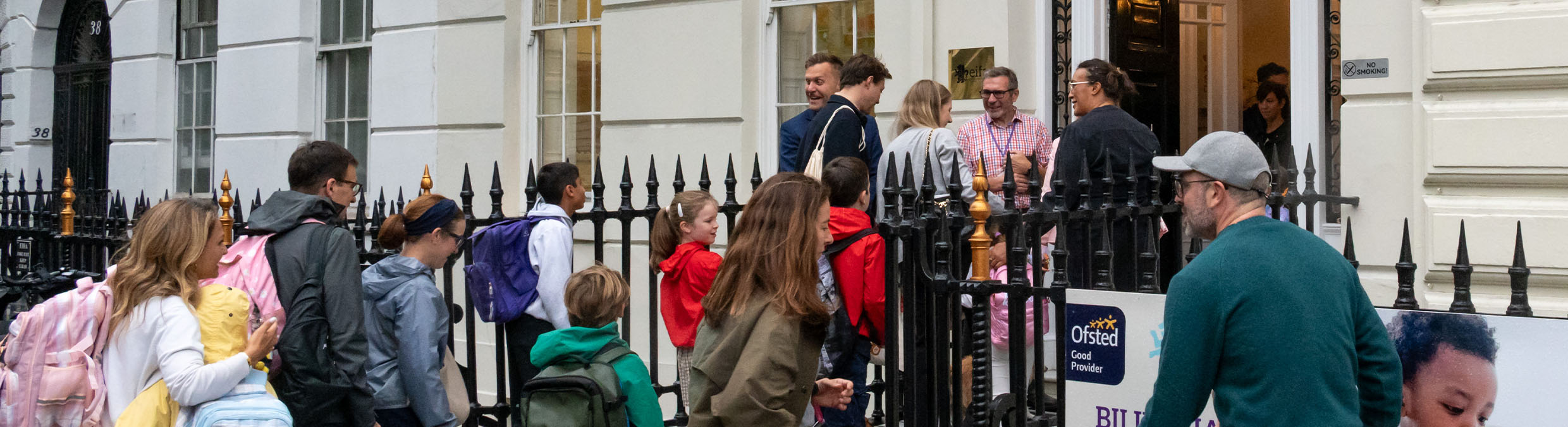 EIFA A group of parents and children with backpacks, including Year 5 and 6 students heading to Lyon, walk toward the entrance of a city building, where several adults are cheerfully greeting them—capturing the bustle of a lively school drop-off.