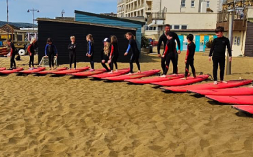 EIFA A group of children in wetsuits stand on pink surfboards arranged in a semi-circle on sandy beach, reflecting EIFA’s commitment to supporting the local community, with beach huts, buildings, and the sea in the background.
