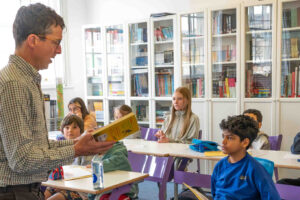 EIFA Un enseignant lit à voix haute à un groupe d'élèves attentifs dans une salle de classe lumineuse et bien éclairée, ornée d'étagères. Tandis que des trousses colorées et des bouteilles d'eau ornent leurs bureaux, ils sont transportés dans un voyage imaginaire, semblable à une visite à Greenwich.