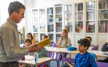 EIFA Un enseignant lit à voix haute à un groupe d'élèves attentifs dans une salle de classe lumineuse et bien éclairée, ornée d'étagères. Tandis que des trousses colorées et des bouteilles d'eau ornent leurs bureaux, ils sont transportés dans un voyage imaginaire, semblable à une visite à Greenwich.