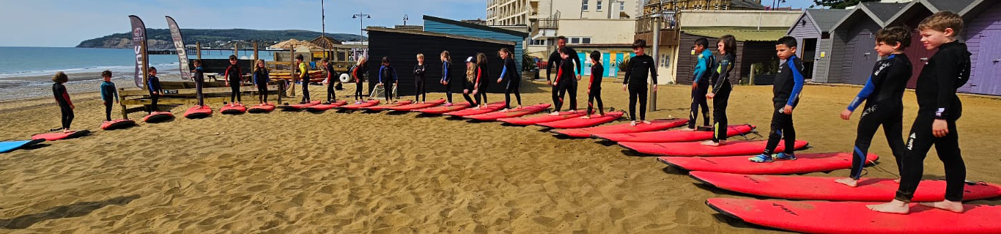 EIFA A group of children in wetsuits stand on pink surfboards arranged in a semi-circle on sandy beach, reflecting EIFA’s commitment to supporting the local community, with beach huts, buildings, and the sea in the background.
