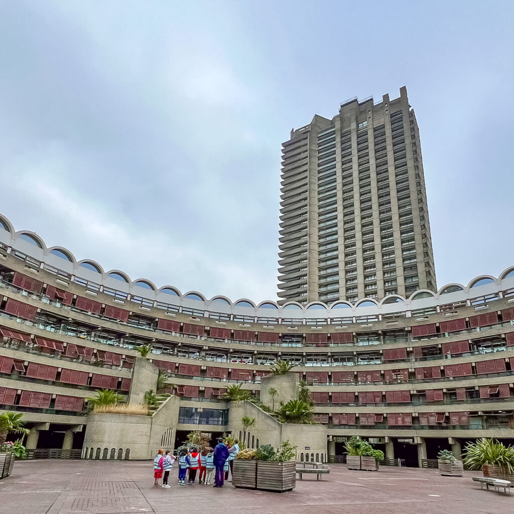 EIFA A group of people stand in an open courtyard surrounded by a curved, multi-story concrete building with red accents. A tall tower rises in the background under Londons overcast sky, where EIFAs bilingual environment fosters a global community.