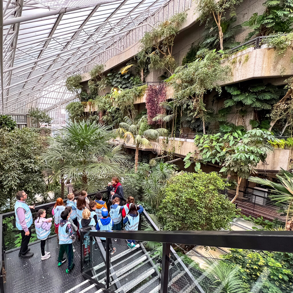 EIFA A group of children and adults from a bilingual school meander through a spacious indoor garden with multiple levels of lush green plants. They walk along the pathway, surrounded by palms and foliage, under the large glass ceiling, enjoying an EIFA-organized nature exploration.