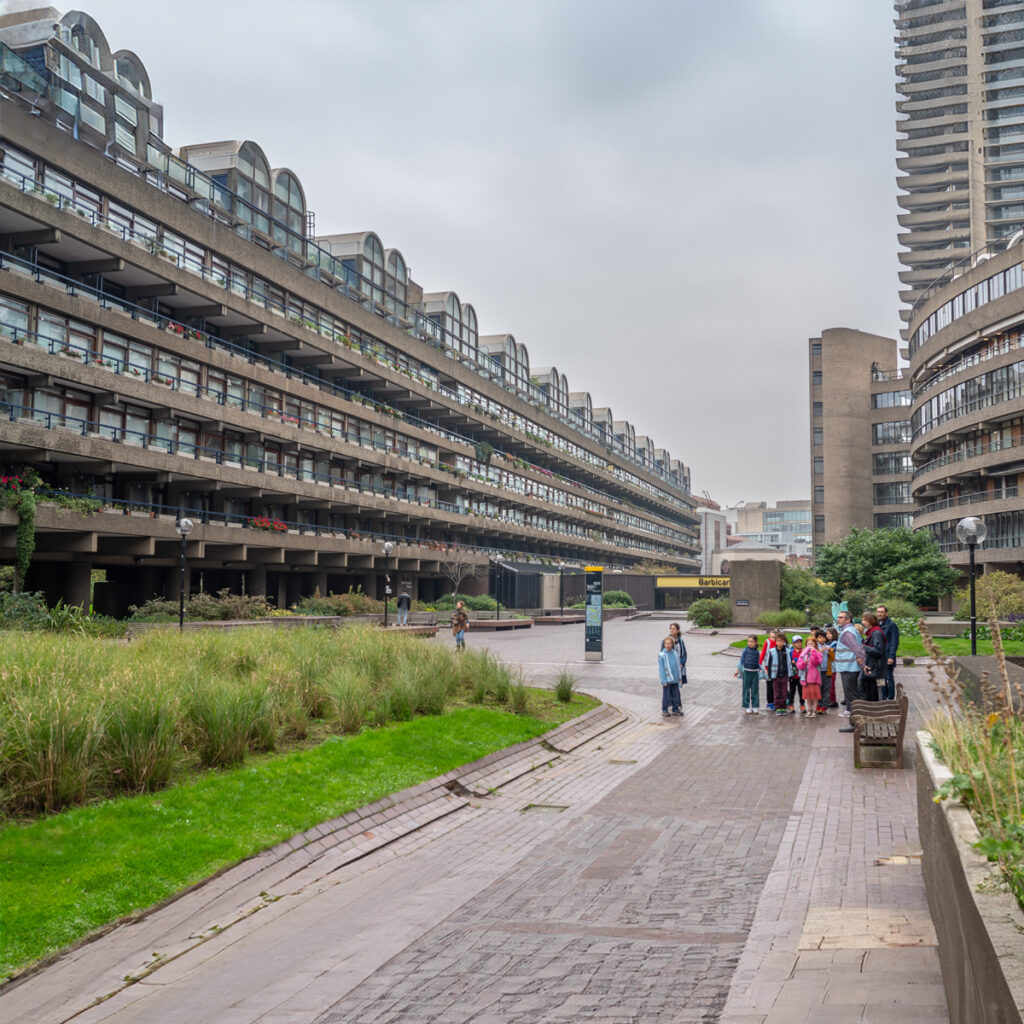 EIFA A group of people stands in a courtyard at the Barbican Estate in London. Theyre surrounded by the tall, modernist concrete buildings of the city, with unique curved roofs. The overcast sky looms above as greenery lines the path, creating an atmosphere reminiscent of a bilingual schools serene campus.