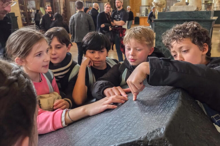 EIFA Un groupe d'enfants examine attentivement et touche des sculptures anciennes sur un objet en pierre dans un musée, avec d'autres visiteurs visibles à l'arrière-plan.