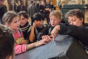 EIFA A group of children closely examine and touch the Rosetta Stone in a museum, with a marble statue and other visitors visible in the background. EIFA A group of children closely examine and touch the Rosetta Stone in a museum, with a marble statue and other visitors visible in the background.