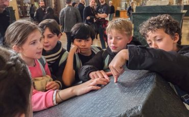 EIFA A group of children closely examine and touch the Rosetta Stone in a museum, with a marble statue and other visitors visible in the background.
