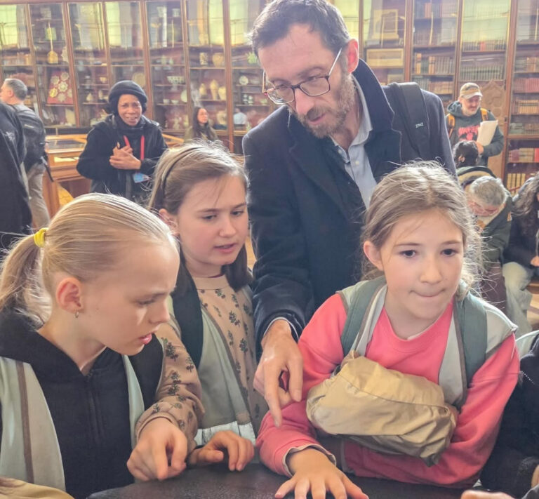 EIFA A man guides three young girls as they closely examine an object on a table in a library or museum, surrounded by bookshelves and other visitors in the background.
