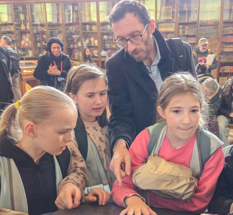 EIFA A bearded man points at something on a table while three young girls watch closely. They are in a library with shelves of books in the background, and other people are visible behind them.