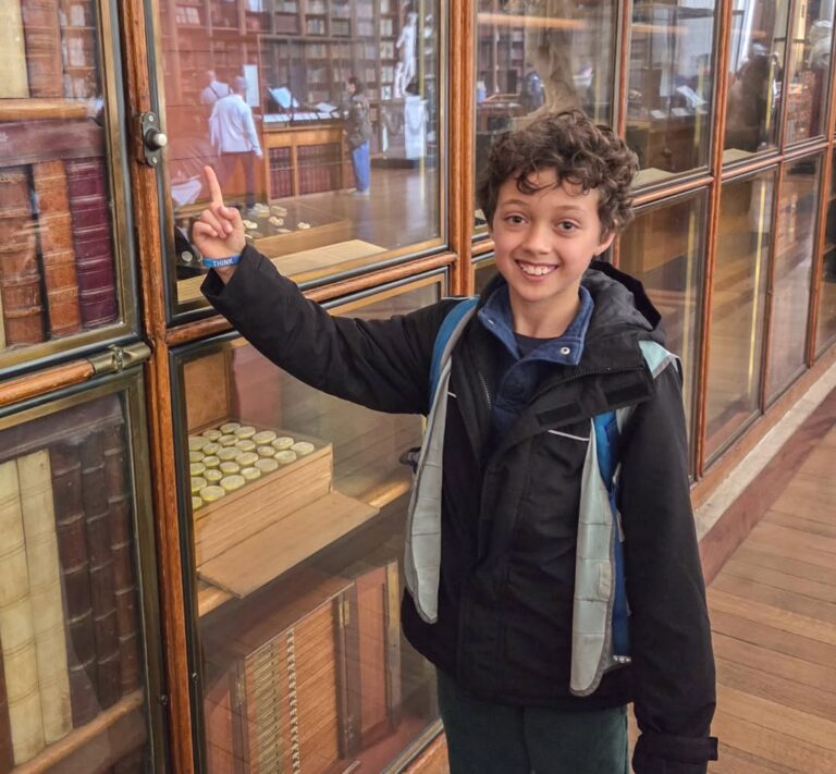 EIFA A smiling child with curly hair and a backpack points at a glass display case filled with old books in a library. More people and shelves of books are visible in the background.