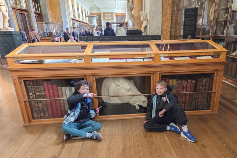 EIFA Deux enfants sont assis par terre devant une vitrine de musée contenant de grands livres anciens et une tête de cheval sculptée. Derrière eux, d'autres visiteurs traversent la galerie.