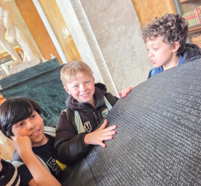 EIFA Trois enfants sourient et touchent une grande pierre noire couverte d'écritures anciennes, exposée dans un musée aux murs de marbre et aux statues en arrière-plan.
