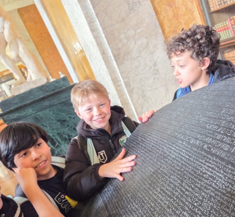 EIFA Three children smile and gather around a large replica of the Rosetta Stone in a museum, looking at the ancient inscriptions. Shelves of books and a marble statue are visible in the background.