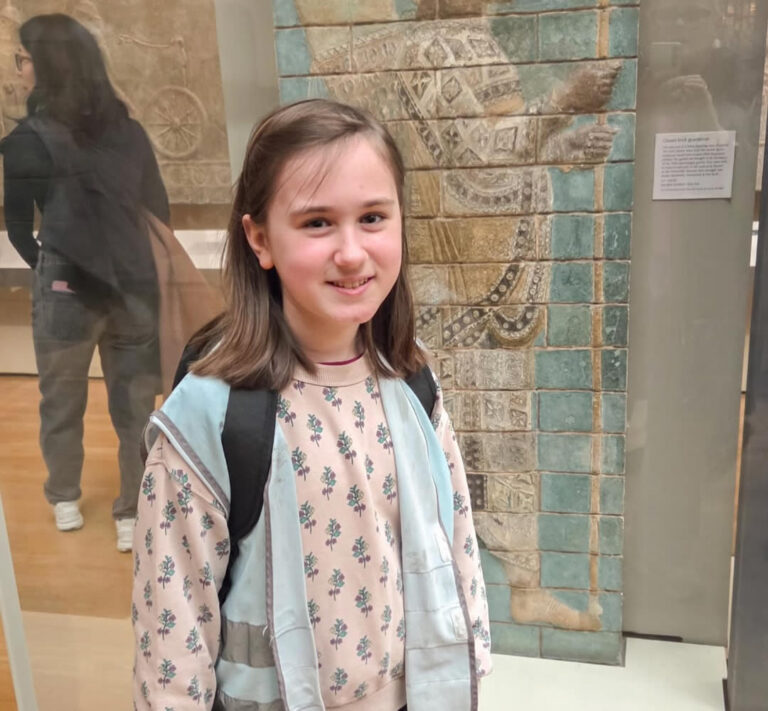 EIFA Une jeune fille aux cheveux bruns, portant une chemise à fleurs, un gilet bleu clair et un sac à dos, sourit devant un ancien relief en pierre dans un musée. Une autre personne est visible à l'arrière-plan.